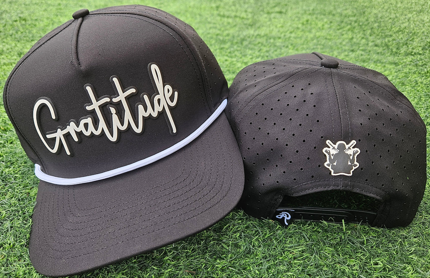 Two black baseball caps on grass, one with 'Gratitude' text and the other with a logo.