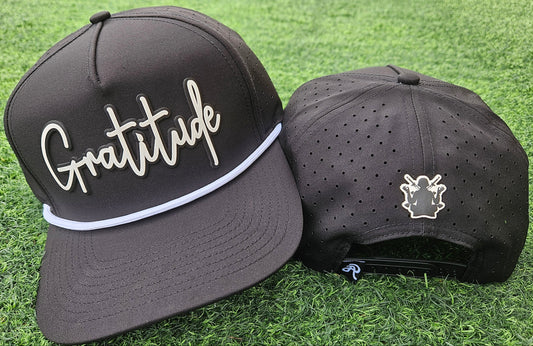 Two black baseball caps on grass, one with 'Gratitude' text and the other with a logo.